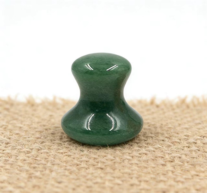 May include: A green jade massage tool with a rounded top and base, connected by a narrower midsection. The polished stone reflects light, and the tool sits on a textured, tan surface. The shape is ergonomic and designed for massage.