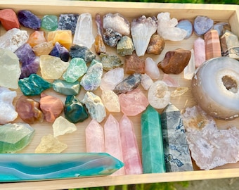 May include: A wooden tray filled with a variety of colorful crystals and gemstones. The collection includes amethyst, rose quartz, clear quartz, and other polished and raw stones in various shapes and sizes. A large geode is also present.