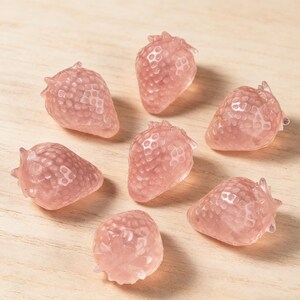 May include: Seven pink strawberry-shaped decorative objects. Each strawberry has a textured surface and a small green stem. The objects are arranged on a light-colored wooden surface.