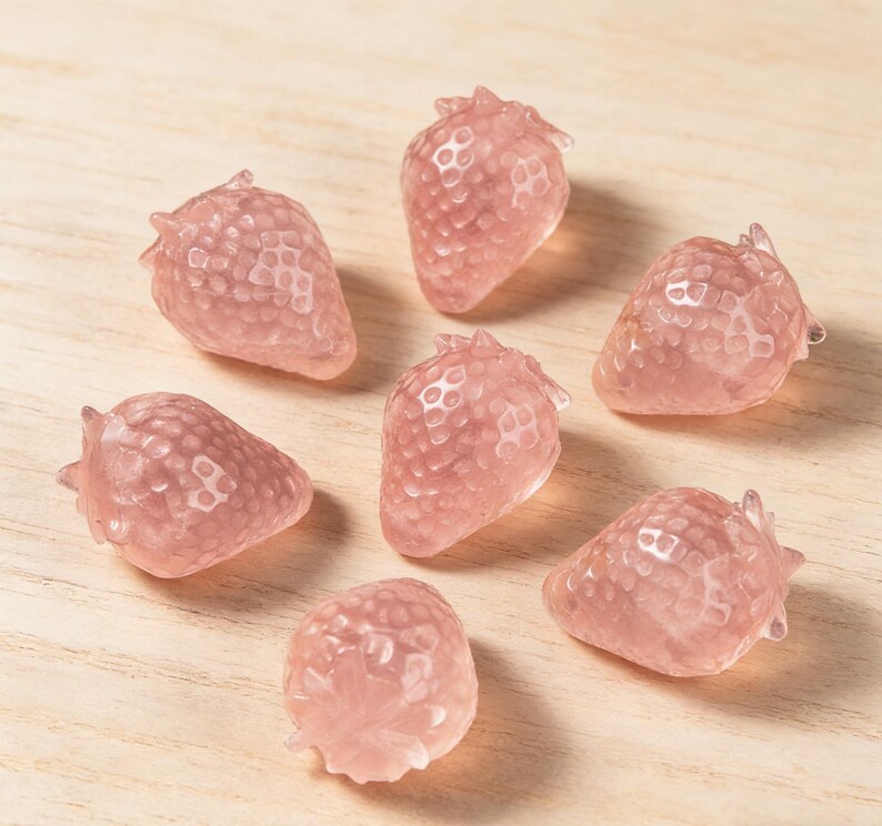 May include: Seven pink strawberry-shaped decorative objects. Each strawberry has a textured surface and a small green stem. The objects are arranged on a light-colored wooden surface.
