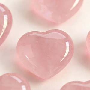 May include: Several polished rose quartz hearts in varying sizes are arranged against a light background. The hearts have a soft pink hue and a smooth, glossy surface, reflecting light. The image is a close-up, highlighting the natural beauty of the stone.