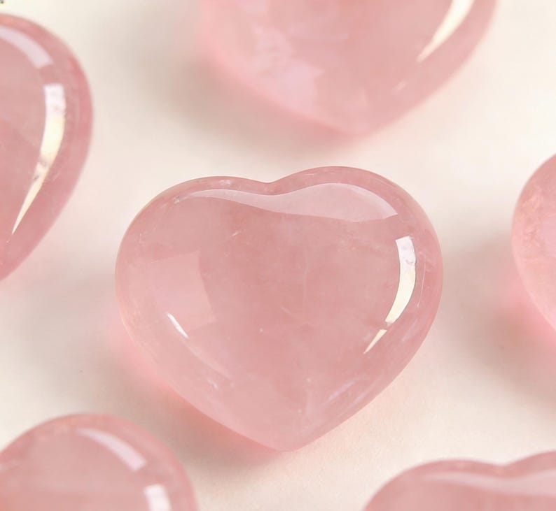 May include: Several polished rose quartz hearts in varying sizes are arranged against a light background. The hearts have a soft pink hue and a smooth, glossy surface, reflecting light. The image is a close-up, highlighting the natural beauty of the stone.