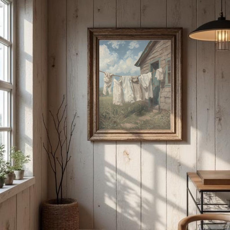 May include: A framed painting showing white laundry hanging on a washing line in front of a weathered wooden building. The scene is set in a field under a blue sky with fluffy clouds. The artwork has a vintage, rustic aesthetic.