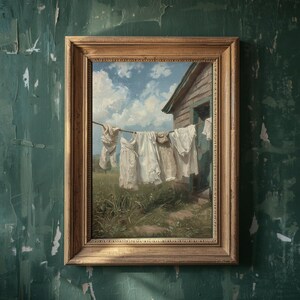 May include: A framed painting showing white laundry hanging on a washing line in front of a small wooden building. The sky is blue with fluffy white clouds. The painting is in a gold-coloured frame.
