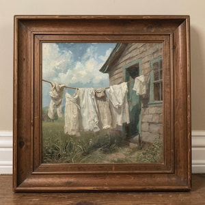 May include: A framed painting showing white laundry hanging on a washing line. The clothes are blowing in the wind in front of a small house with a blue door and window. The painting is set in a grassy field under a blue sky with clouds.