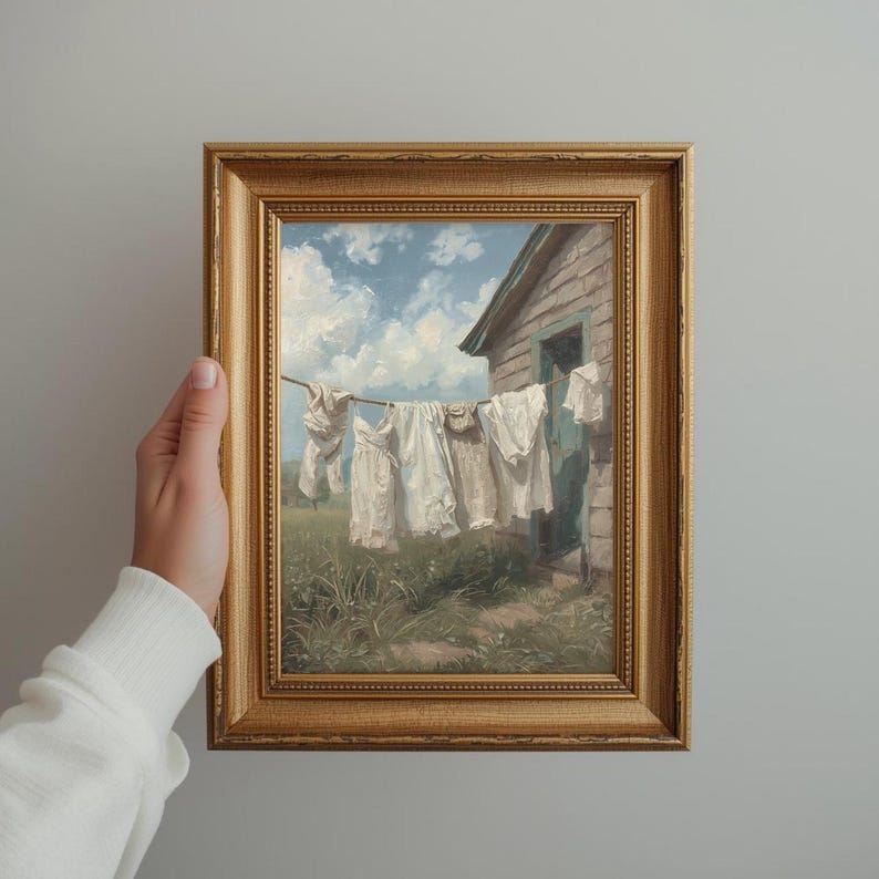 May include: A framed painting shows white laundry hanging on a washing line in front of a weathered building. The artwork features a blue sky with fluffy clouds and green grass. The frame is gold.