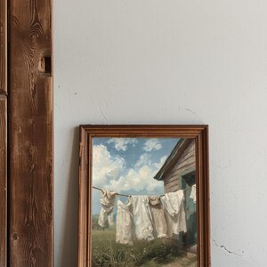 May include: A framed painting showing white laundry hanging on a washing line against a blue sky with clouds. The artwork is in a wooden frame, leaning against a white wall and a wooden door frame.