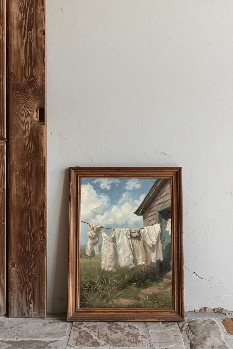 May include: A framed painting showing white laundry hanging on a washing line against a blue sky with clouds. The artwork is in a wooden frame, leaning against a white wall and a wooden door frame.