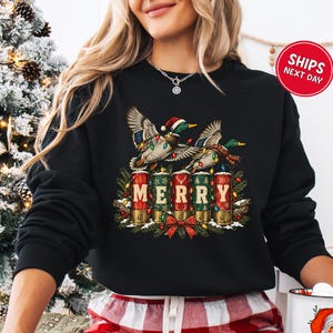 May include: A black crewneck sweatshirt featuring a graphic of two mallard ducks wearing Santa hats and wrapped in Christmas lights. Below the ducks are five shotgun shells arranged to spell MERRY, decorated with pine branches and a red bow.