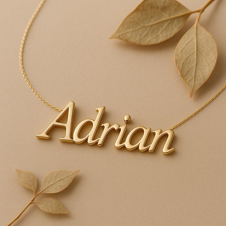 May include: A gold-colored necklace with the name "Adrian" in a cursive font. The necklace is on a delicate chain, and the name is the focal point. The background is a neutral beige color, with dried leaves.