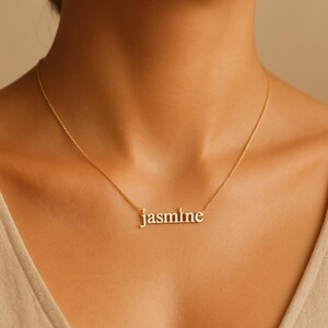 May include: A gold-colored necklace with the name "Jasmine" spelled out in lowercase letters. The necklace is worn around the neck and hangs against a person's skin.