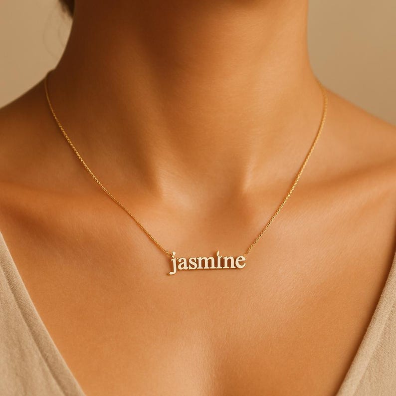May include: A gold-colored necklace with the name "Jasmine" spelled out in lowercase letters. The necklace is worn around the neck and hangs against a person's skin.
