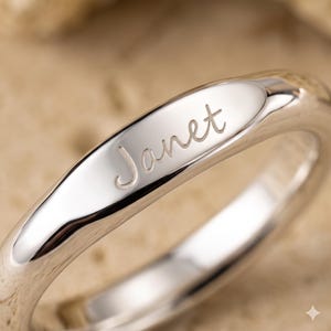 May include: A polished silver-toned ring featuring a smooth, rounded band with the name Janet engraved in a cursive script on the front surface. The metal has a reflective finish, highlighting the depth and clarity of the engraved lettering.