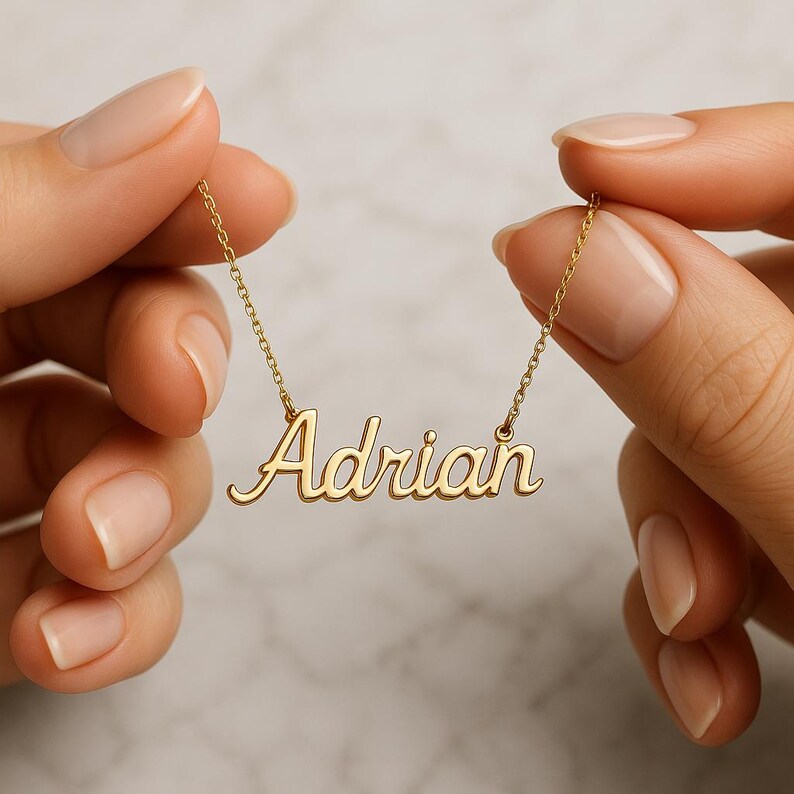 May include: A gold-colored necklace with the name "Adrian" in cursive script. The necklace is held by two hands with manicured nails, showcasing the delicate chain and pendant design. The background is blurred, focusing attention on the jewelry.
