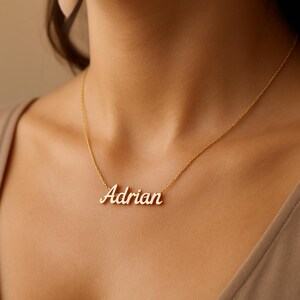 May include: A gold-colored necklace with the name "Adrian" in a cursive font. The pendant is suspended from a delicate chain. The necklace is displayed on a person's neck.