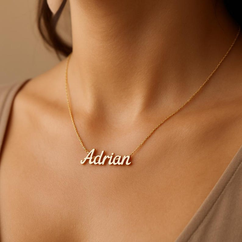 May include: A gold-colored necklace with the name "Adrian" in a cursive font. The pendant is suspended from a delicate chain. The necklace is displayed on a person's neck.