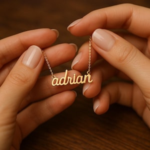 May include: A gold-colored necklace with the name "adrian" in a cursive font. The necklace is held between two hands with manicured nails, set against a blurred background.