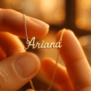 May include: A gold-colored necklace with the name "Ariana" in a cursive font. The necklace is held between two sets of fingers, with the chain visible. The background is blurred, with warm tones.