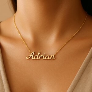 May include: A gold-colored necklace with the name "Adrian" in a cursive font. The pendant hangs from a delicate chain, resting against a person's skin. The necklace is a personalized jewelry item.