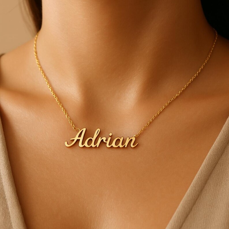 May include: A gold-colored necklace with the name "Adrian" in a cursive font. The pendant hangs from a delicate chain, resting against a person's skin. The necklace is a personalized jewelry item.