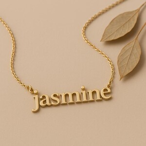 May include: A gold-colored necklace with the name "Jasmine" spelled out in block letters. The necklace is on a delicate chain. Two dried leaves are in the background, set against a neutral backdrop.