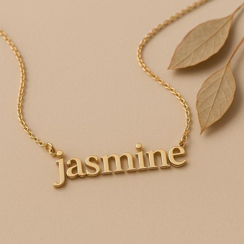 May include: A gold-colored necklace with the name "Jasmine" spelled out in block letters. The necklace is on a delicate chain. Two dried leaves are in the background, set against a neutral backdrop.