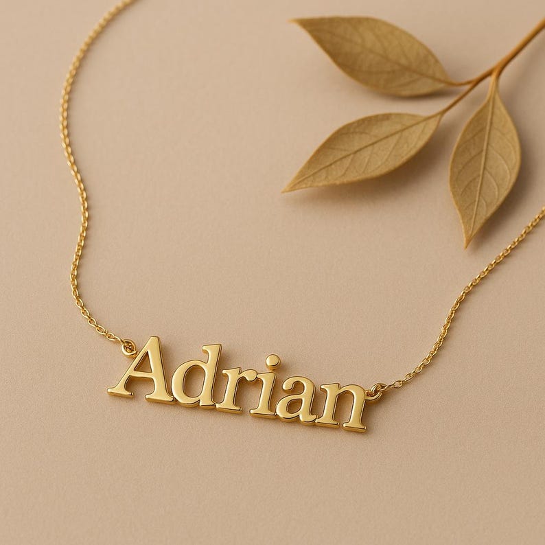 May include: A gold-colored necklace with the name "Adrian" in block letters. The necklace is on a delicate chain. The background is a neutral beige color with a sprig of dried leaves.