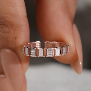 May include: A rose gold band ring featuring an alternating pattern of rectangular white enamel segments and clear baguette-cut stones. The ring has an open back design, allowing for adjustable sizing, and is constructed with a series of linked metal segments.