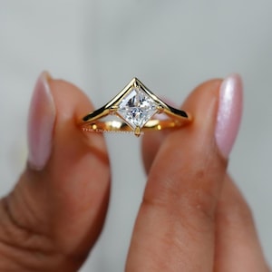 May include: A gold ring with a princess-cut diamond. The diamond is set in a V-shaped design, and the band is a simple, polished gold. The ring is held between two fingers.