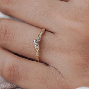 May include: A gold-colored ring with a leaf and vine design. The ring features several small, clear gemstones. The ring is curved and sits on a finger. The ring is a piece of jewelry.
