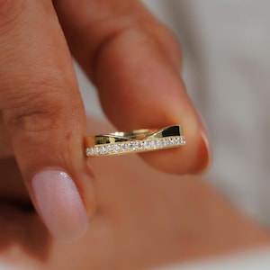 Puede incluir: Un anillo dorado con un diseño ondulado. El anillo está engastado con pequeñas piedras preciosas transparentes a lo largo del borde superior. El anillo se sostiene entre los dedos, con un fondo borroso.