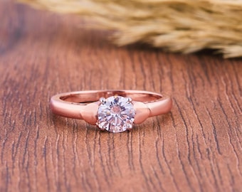 May include: Rose gold engagement ring with a large, round, clear gemstone. The ring has a simple band with decorative accents on either side of the gemstone. The ring is set on a wooden surface.