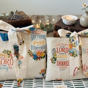 May include: Cream-colored tote bags with floral and scripture designs. One bag reads "Sing to the Lord a new song," another says "Worry about anything, pray about everything," and the third says "Give thanks to the Lord." Each bag has a matching ribbon.