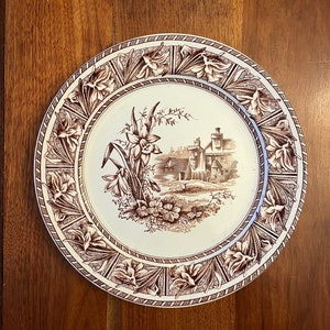 May include: A brown and white ceramic plate with a floral design. The plate features a cottage scene with a daffodil flower in the foreground. The border of the plate is decorated with a repeating pattern of daffodils.