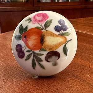 May include: A white ceramic knob with a painted design of fruit and flowers. The knob features a pear, apple, mushroom, and a pink flower.