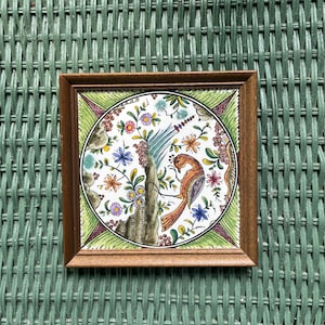 May include: A framed ceramic tile with a colorful design of a bird surrounded by flowers and leaves. The tile is set against a green wicker background.