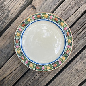 May include: A white ceramic plate with a blue and yellow rim. The rim is decorated with a repeating pattern of green leaves, pink flowers, and brown acorns.