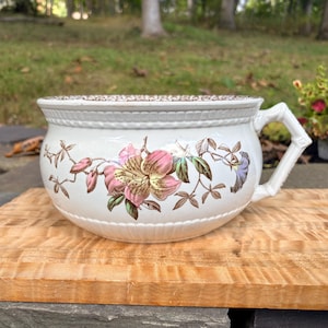 May include: A white ceramic chamber pot featuring a floral pattern with pink and brown blossoms and green leaves. The vessel has a textured rim, a sturdy handle, and a brown patterned lid. The body is rounded with a smooth, glossy finish.