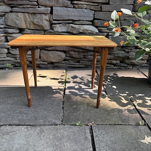 May include: A rectangular wooden side table with a smooth, light brown finish. The table features four tapered legs with subtle ringed detailing near the feet and thin, dark metal support braces connecting the legs to the underside of the top.