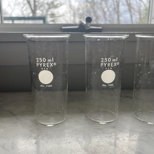 May include: Three clear Pyrex beakers, each labeled "250 ml PYREX® USA" and "No. 1140". The beakers are cylindrical with a flat base and are positioned on a light-colored surface near a window.