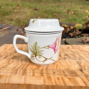 May include: A white ceramic mug with a matching lid, featuring a botanical illustration of a pink morning glory flower with green leaves on the side and a small green leaf detail on the top of the lid, complete with a side handle.