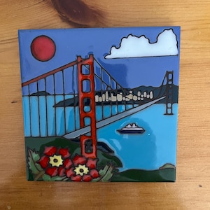 May include: A ceramic tile with a painted image of the Golden Gate Bridge in San Francisco. The bridge is red and the water is blue. There are red flowers in the foreground and a white cloud in the sky.