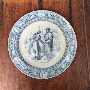 May include: A blue and white ceramic plate with a decorative border featuring a scene of a woman and a man. The scene is surrounded by a border of stylized flowers, birds, and fish. The text "A Piece of Money is a good..." is printed on the plate.