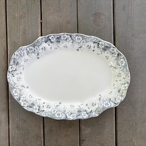 May include: An oval-shaped ceramic platter featuring a scalloped edge and a decorative border of grey and white floral patterns. The center of the platter is a solid, smooth off-white color, while the rim is detailed with intricate botanical illustrations.