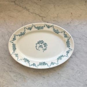 May include: An oval-shaped ceramic serving platter with a cream-colored base. The platter features a decorative border of teal-colored floral garlands and a matching circular floral motif centered in the middle of the dish's smooth surface.