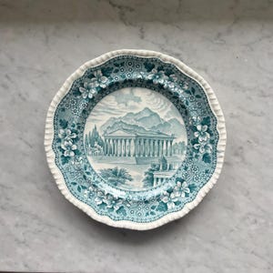 May include: A vintage blue and white porcelain plate with a detailed image of a Greek temple and surrounding landscape. The plate has a scalloped edge and a floral border.