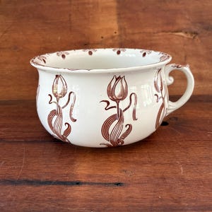 May include: A white ceramic chamber pot featuring a repeating brown tulip pattern around the exterior and a single curved handle. The rim is decorated with a small brown floral motif, and the interior is plain white with a smooth finish.