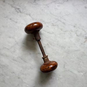 May include: A vintage door knob set featuring two round, polished brown marbled knobs connected by a square metal spindle. The hardware includes a rusted metal collar and a small set screw visible on the neck of one knob, showing signs of age.