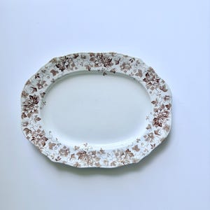May include: An oval ceramic serving platter featuring a white center and a scalloped rim decorated with a brown floral pattern of leaves and blossoms. The intricate botanical design borders the entire edge of the dish in a monochromatic brown tone.