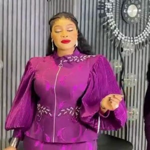 May include: A vibrant purple two-piece outfit consisting of a long-sleeved jacket and a matching skirt. The jacket features a full-length front zip, pleated sleeves, and decorative beaded embellishments on the chest. The fabric has a subtle pattern.